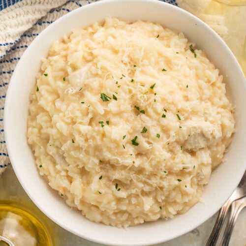A large white bowl of Instant Pot chicken risotto on a table with a blue and white kitchen towel next to a glass of white wine.
