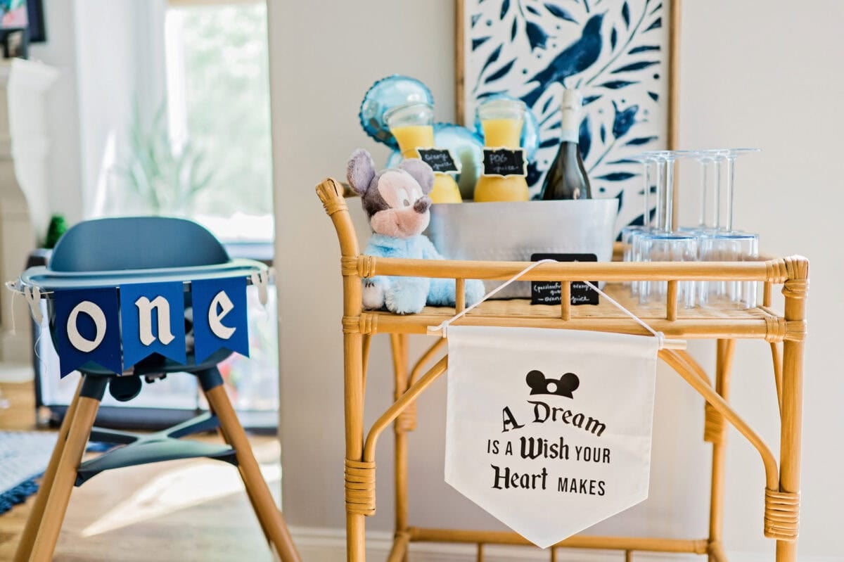 A high chair with a "ONE" banner hanging on the front sits next to a rattan cart decorated as a mimosa bar for a Magic Kingdom birthday party.