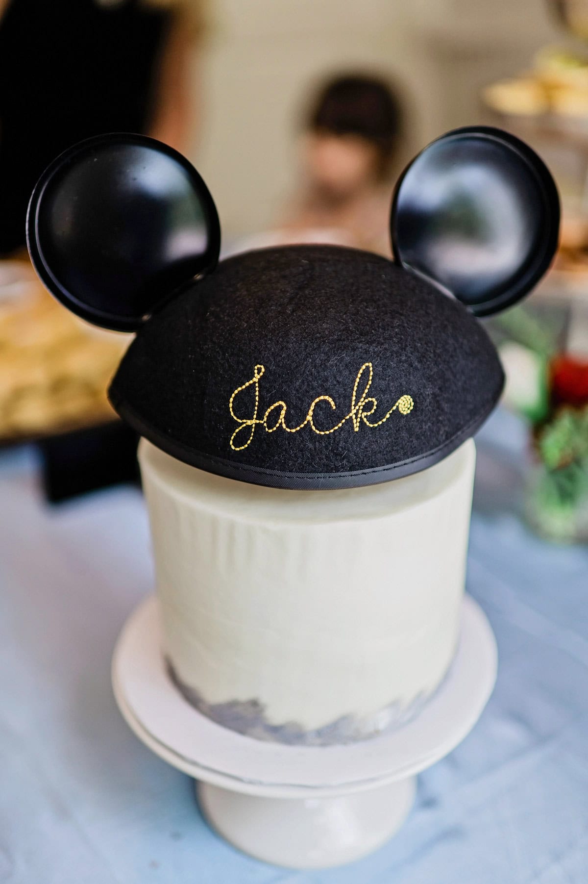 A white round birthday cake topped with a Disneyland classic ear hat. The back of the hat shows cursive gold embroidery spelling out the name "Jack".