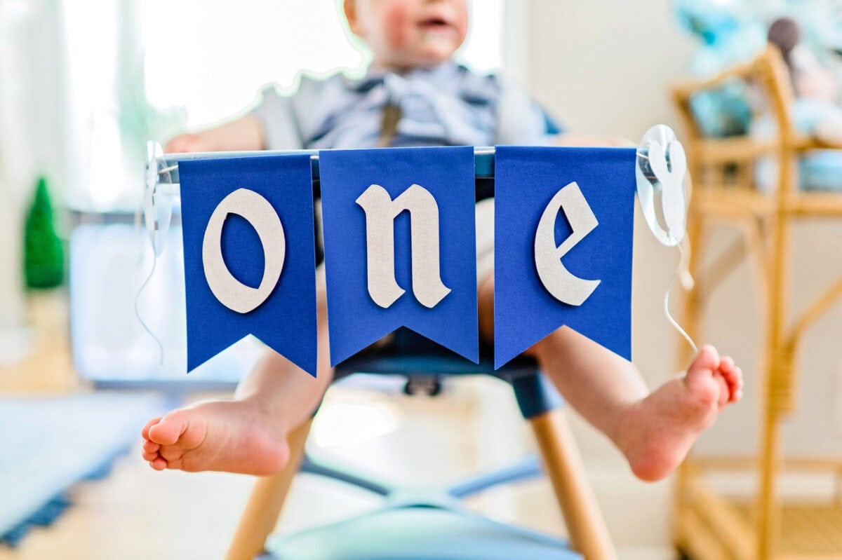 A "ONE" birthday banner hangs on the front of a high chair that a little boy is sitting in.