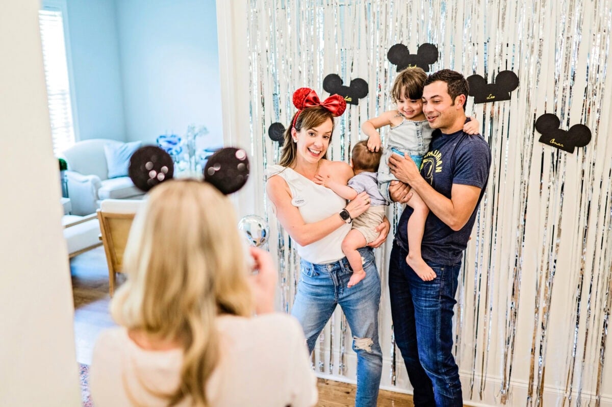 A man and a woman holding two small children pose for a photo in front of silver streamers hanging on a wall.