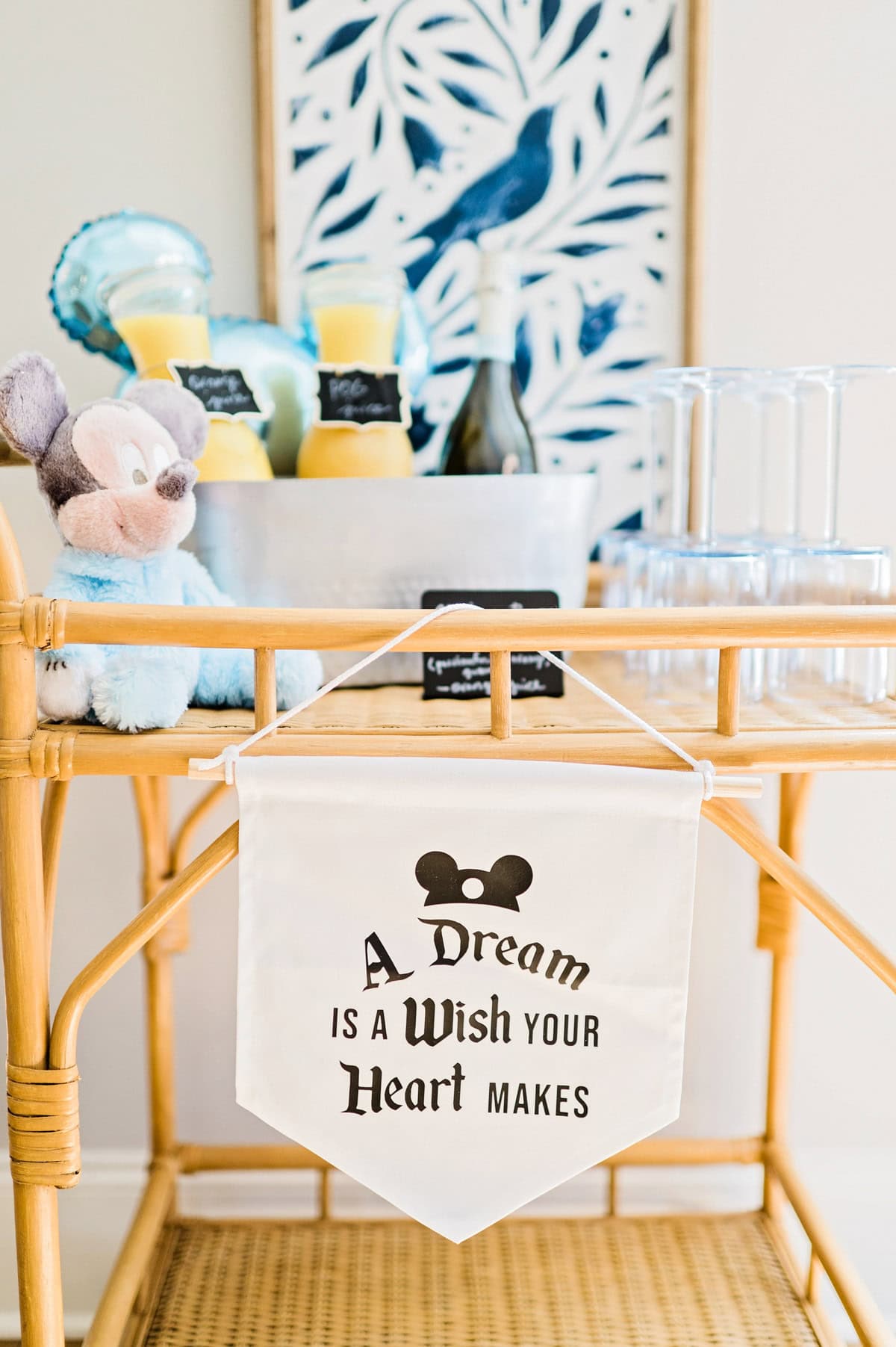 A beverage cart themed for a Magic Kingdom birthday party, with a serve-yourself mimosa bar, decorated with a pastel baby Mickey Mouse plush and a banner that reads "A dream is a wish your heart makes".