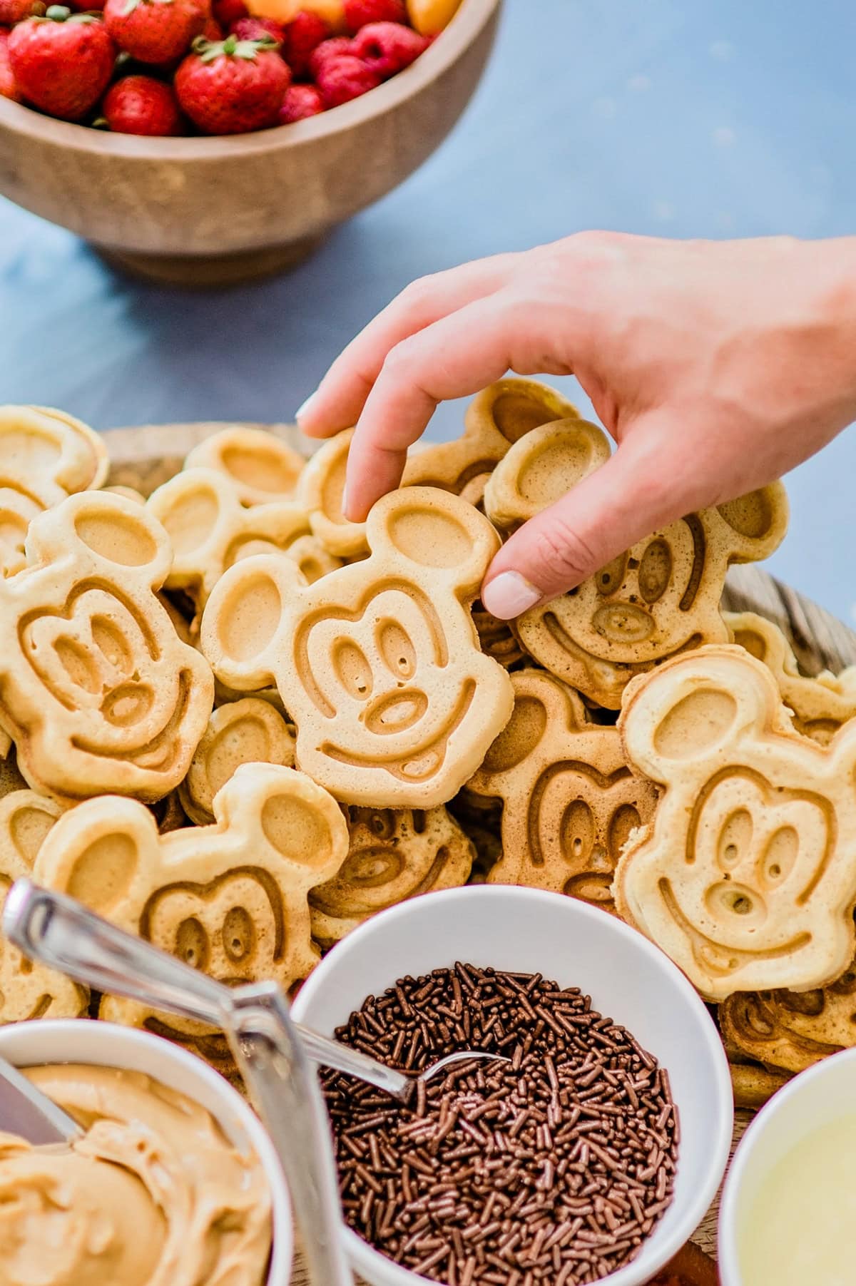 A white woman's hand reaches for a Mickey Mouse shaped waffle on a large tray with waffle toppings.