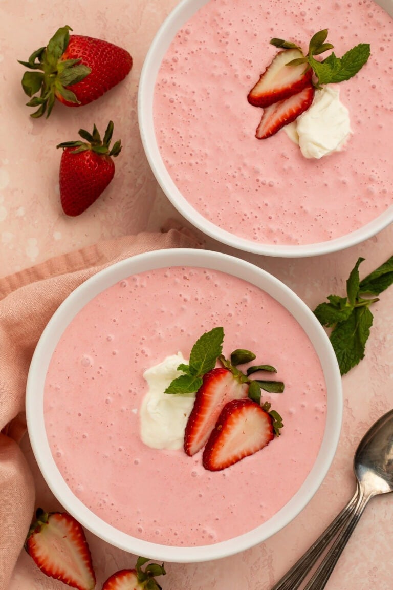 Two bowls of creamy, light pink strawberry soup on a table. The bowls are garnished with sliced strawberries and cream.