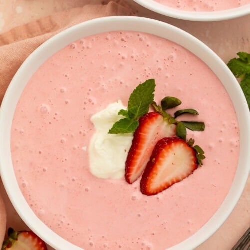 A bowl of creamy, light pink strawberry soup on a table. The soup is garnished with sliced strawberries and cream.