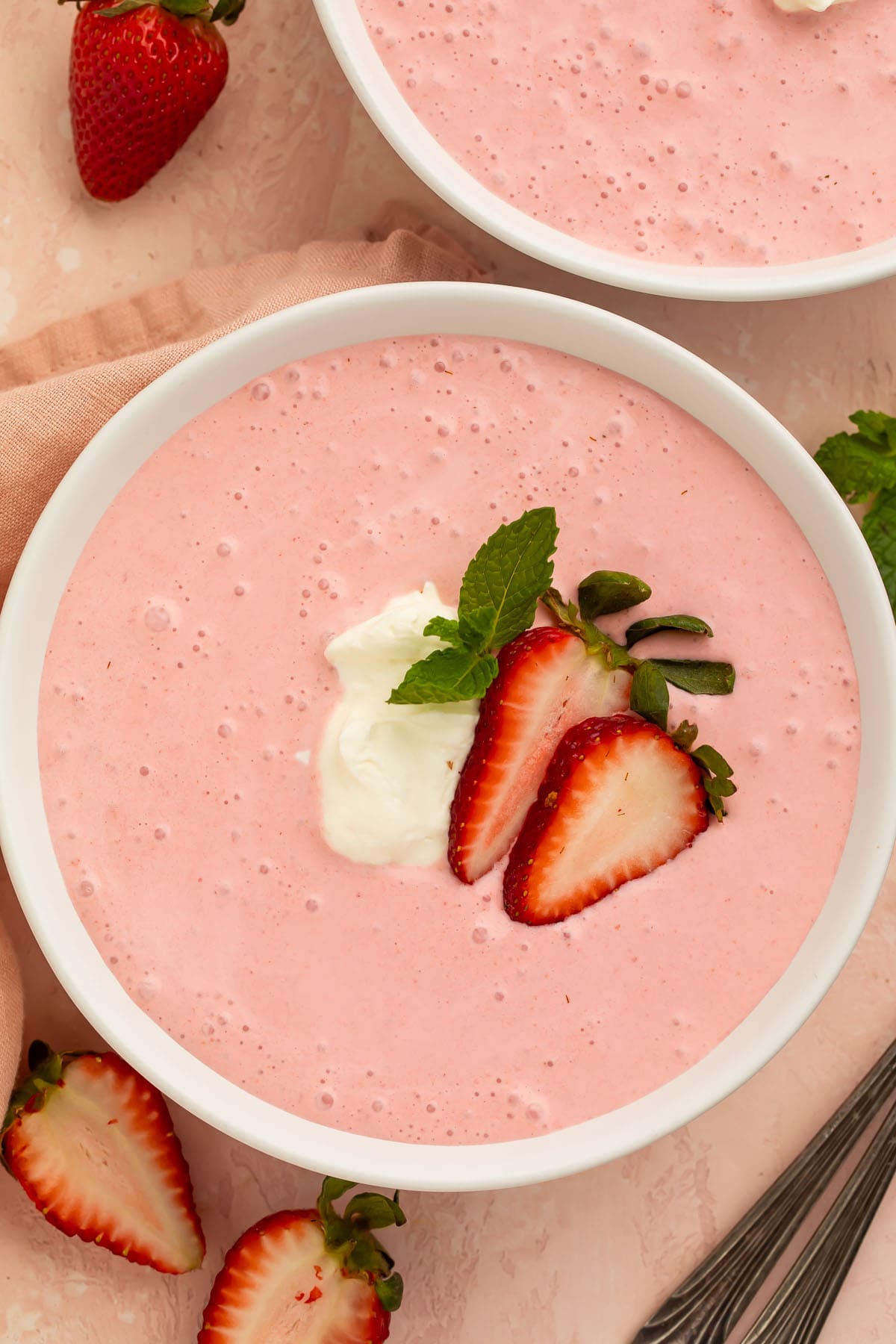 A bowl of creamy, light pink strawberry soup on a table. The soup is garnished with sliced strawberries and cream.
