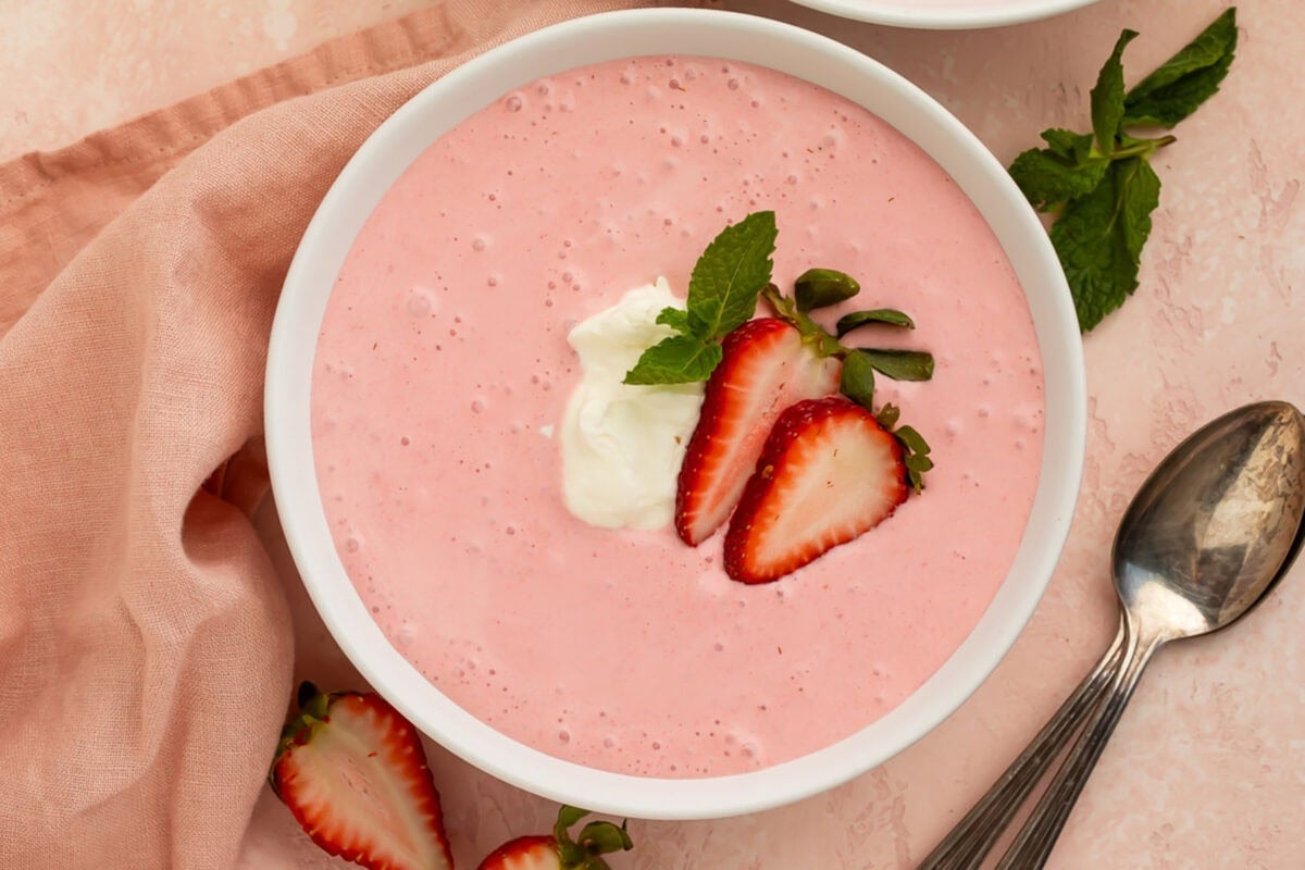 A bowl of creamy, light pink strawberry soup on a table. The soup is garnished with sliced strawberries and cream.