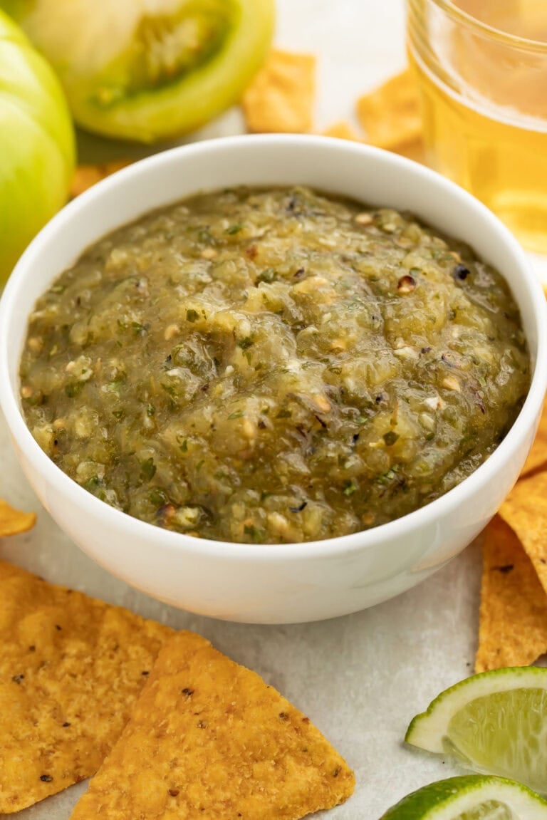 An angled photo of a bowl of green tomato salsa surrounded by white corn tortilla chips.