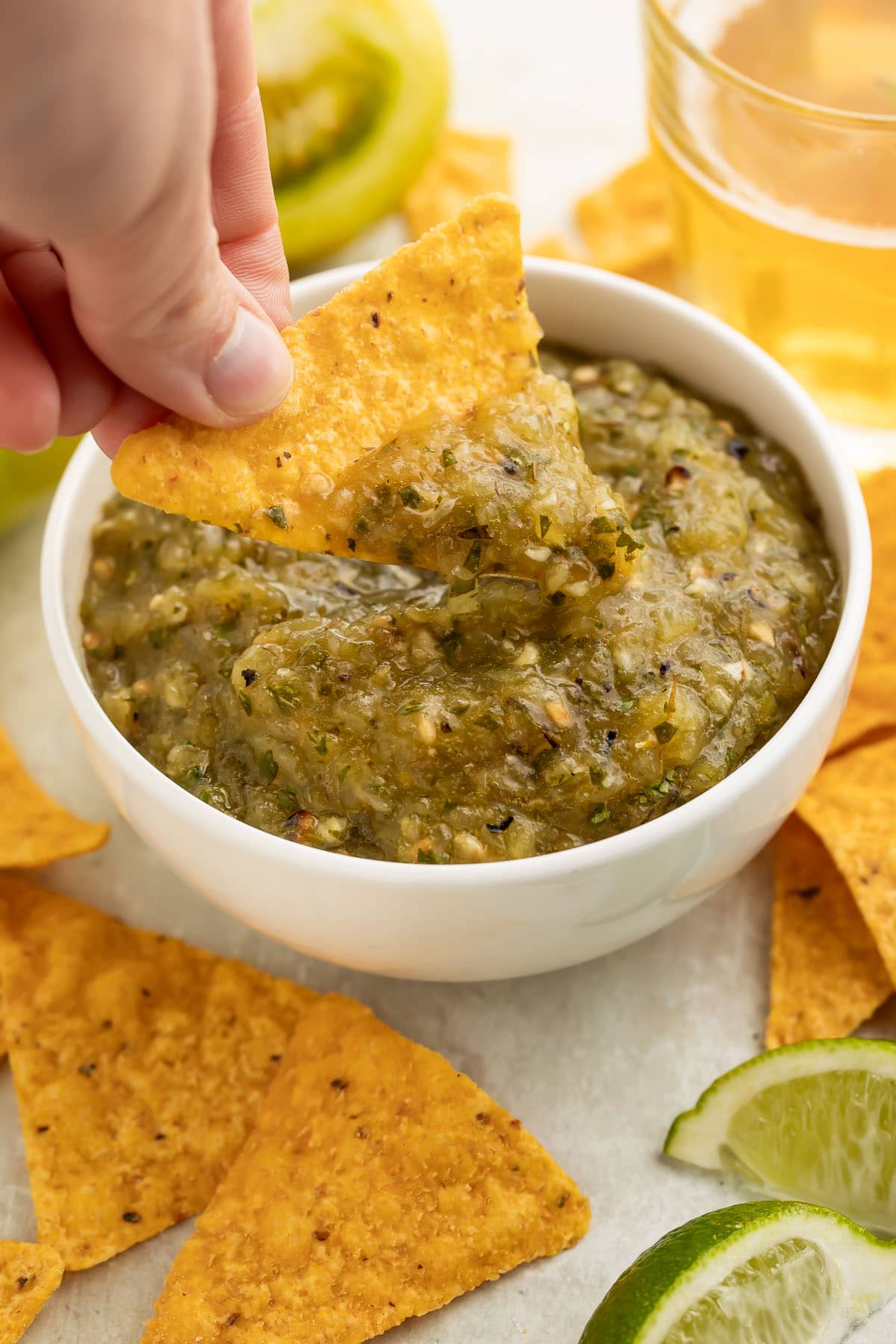 A woman's hand holds a corn tortilla chip as she uses it to scoop green tomato salsa out of a white bowl.