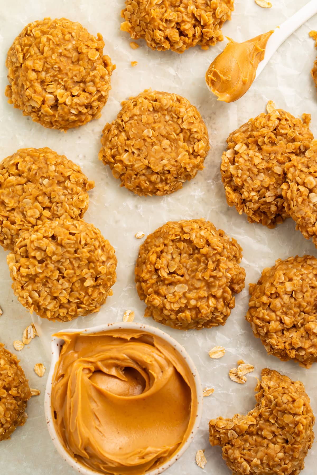 Peanut butter oat cookies on a piece of parchment paper with a measuring cup of smooth peanut butter and a white spoonful of peanut butter.
