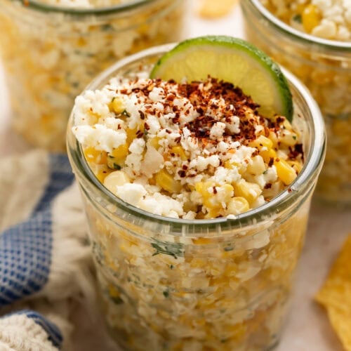 Three glass jars filled with Mexican street corn topped with crumbled cheese, chili powder, and lime wedges
