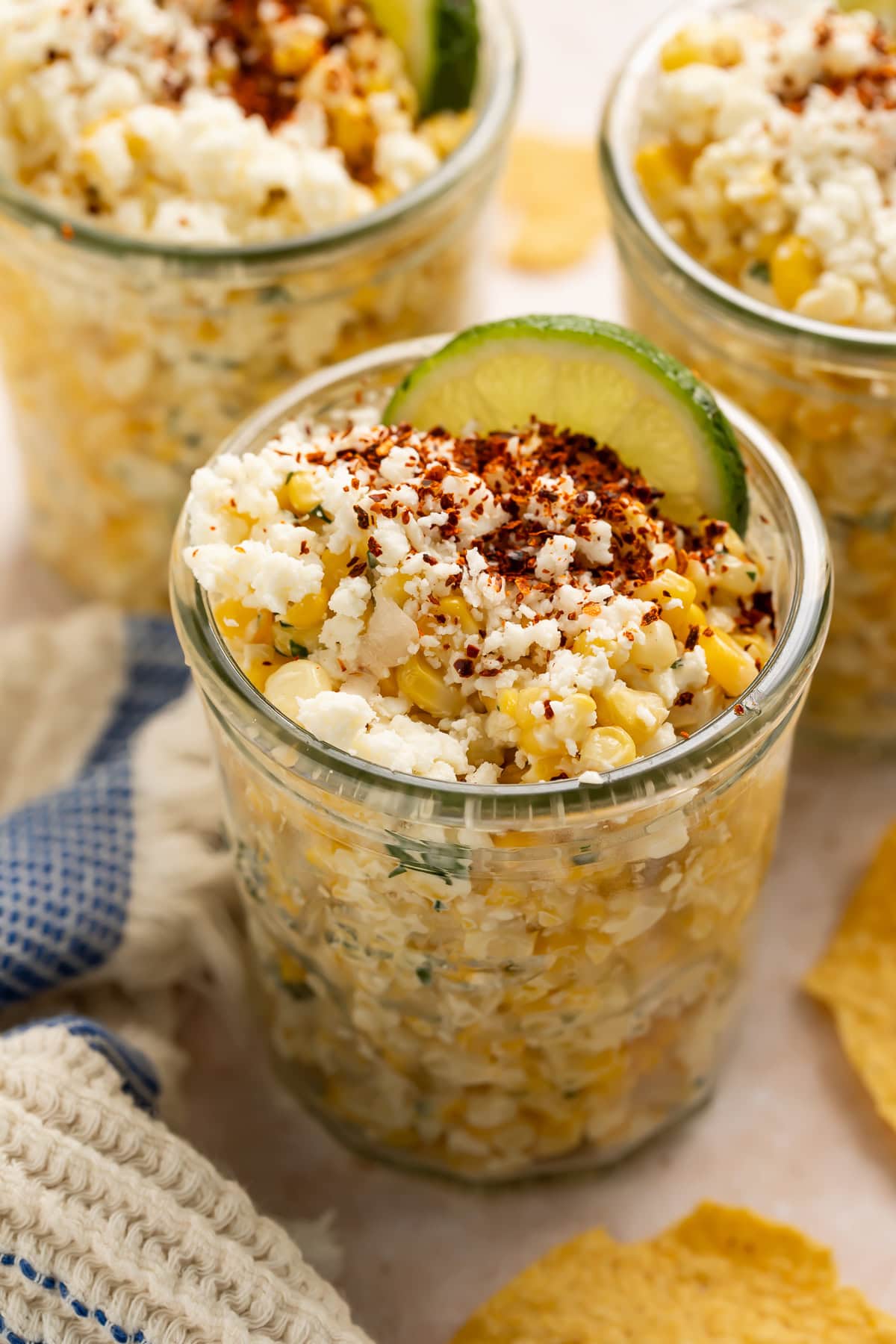 Three glass jars filled with Mexican street corn topped with crumbled cheese, chili powder, and lime wedges