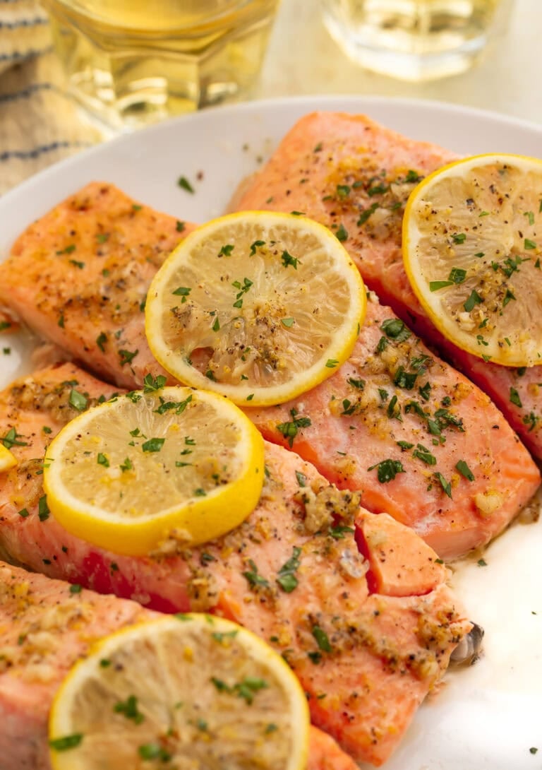 Four baked lemon pepper salmon fillets in white baking dish with lemon slices and fresh parsley
