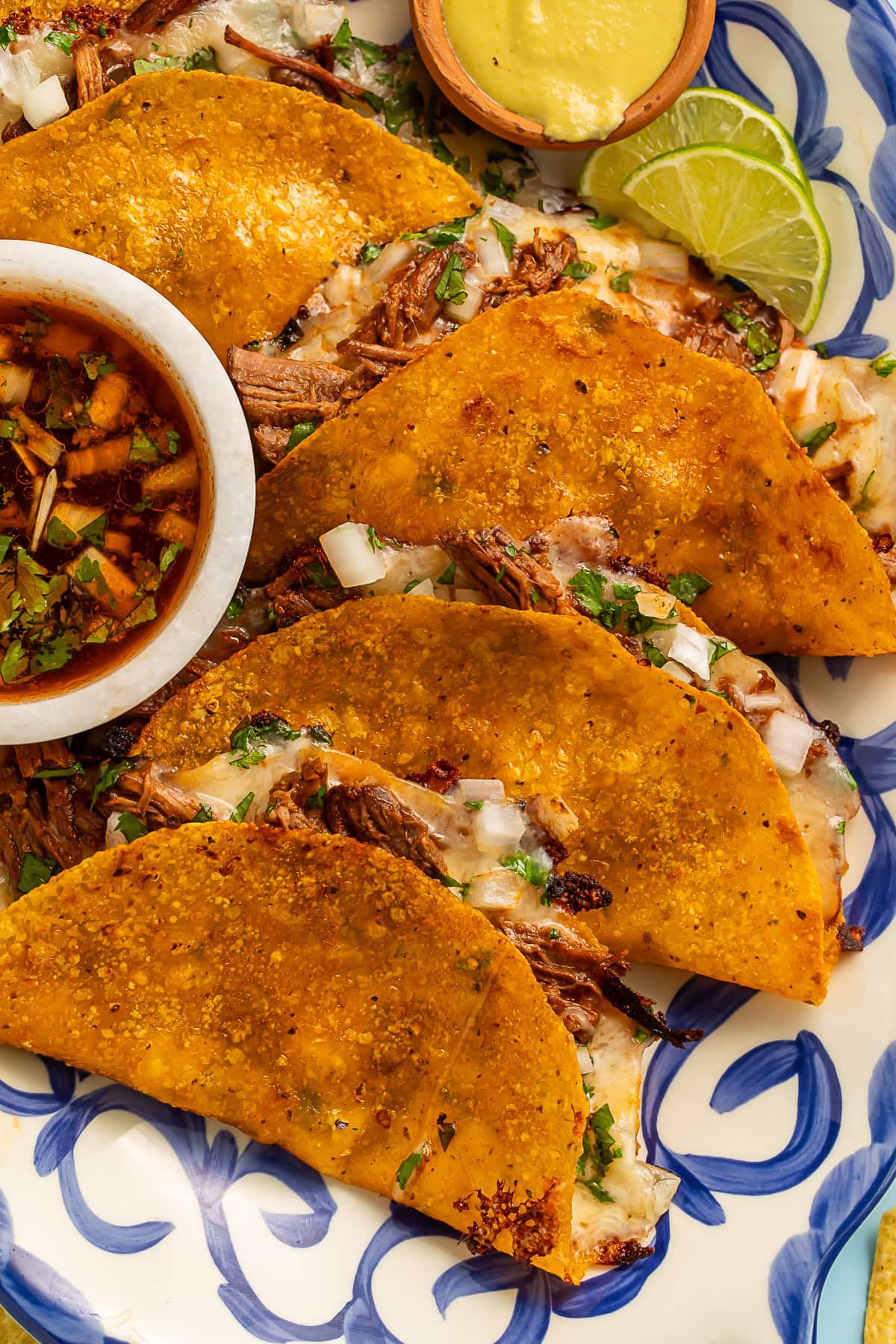 Four golden-brown birria tacos with melted cheese and shredded beef on a blue and white patterned plate, served with consommé for dipping, lime wedges, and garnished with cilantro and diced onions
