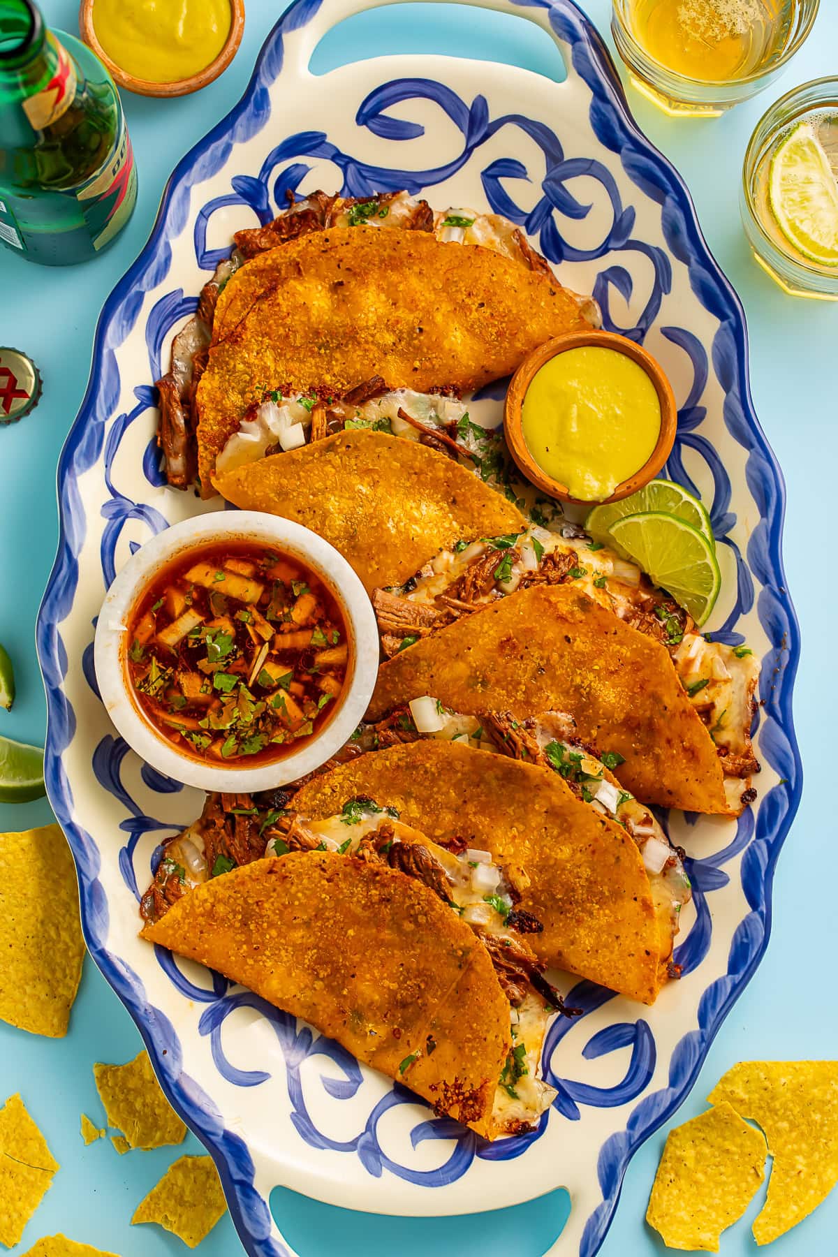 our golden-brown birria tacos with melted cheese and shredded beef on a blue and white patterned plate, served with consommé for dipping, lime wedges, and garnished with cilantro and diced onions