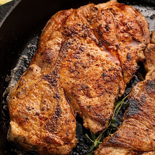 Golden-brown seared pork steaks in black cast iron skillet with fresh thyme sprigs