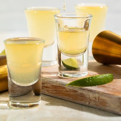 Three green tea shots lined up on bar with lime garnish and cocktail shaker