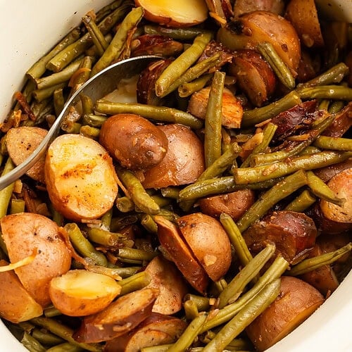 White slow cooker filled with cooked swamp potatoes showing tender red potatoes, green beans, and sliced sausage with serving spoon