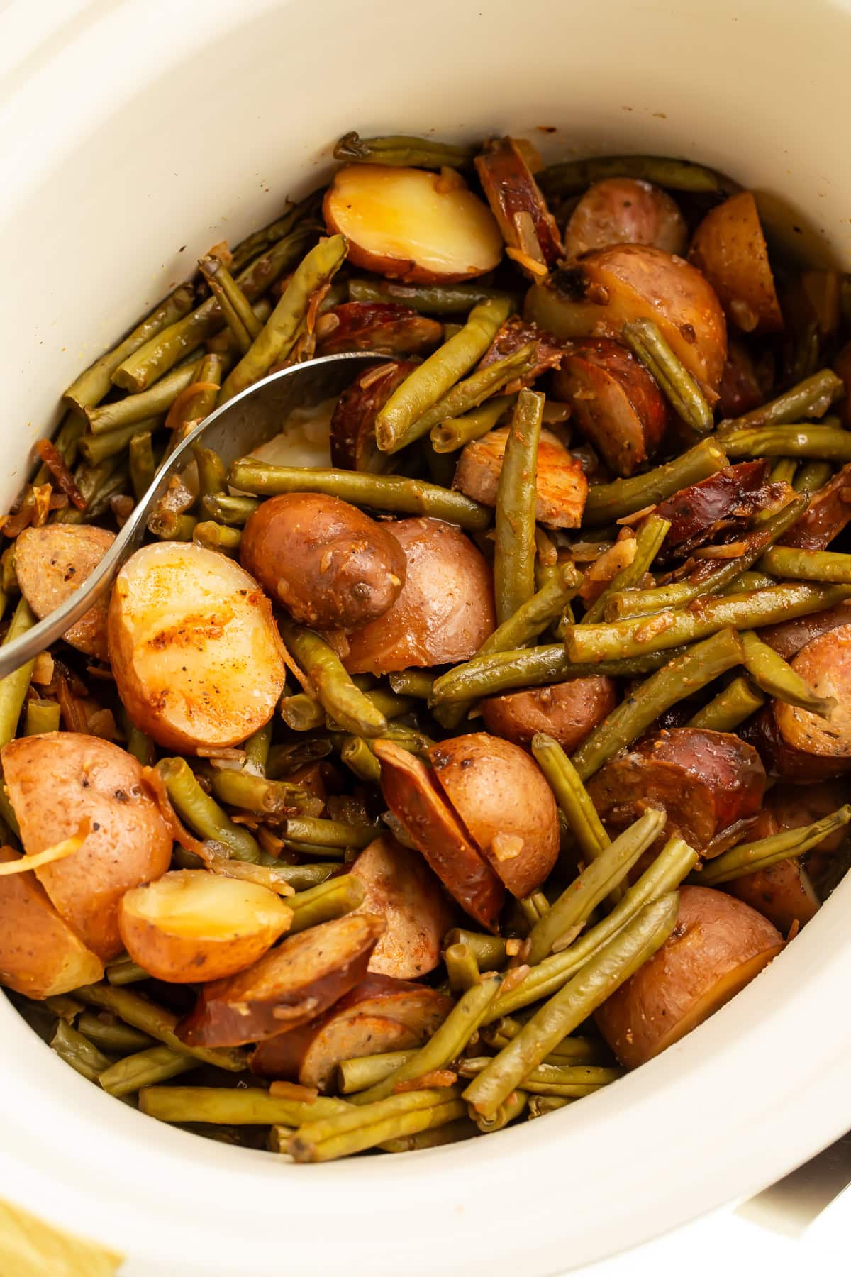 White slow cooker filled with cooked swamp potatoes showing tender red potatoes, green beans, and sliced sausage with serving spoon