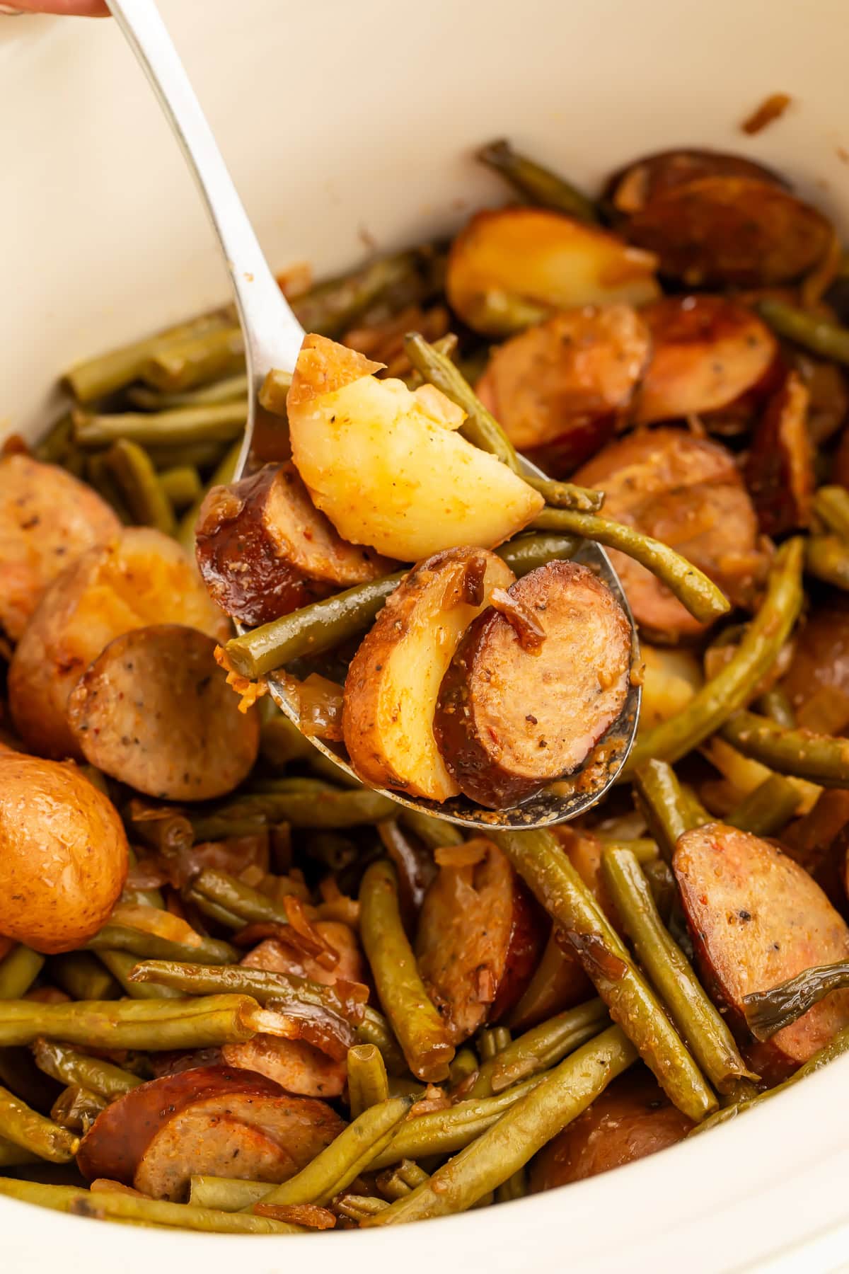 Close-up of serving spoon holding swamp potatoes with golden potato pieces, green beans, and sausage slices