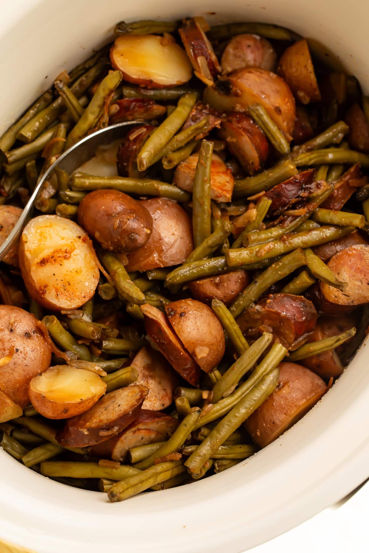 White slow cooker filled with cooked swamp potatoes showing tender red potatoes, green beans, and sliced sausage with serving spoon