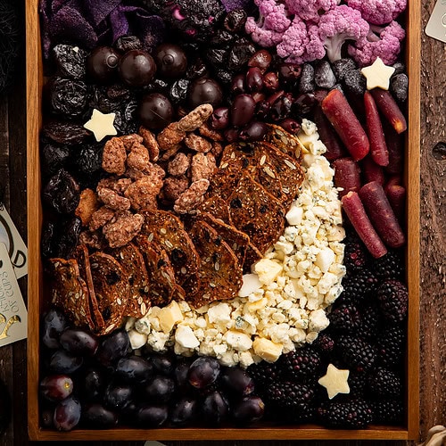 Halloween themed charcuterie board in wooden tray featuring purple and black foods arranged with star cheese cutouts