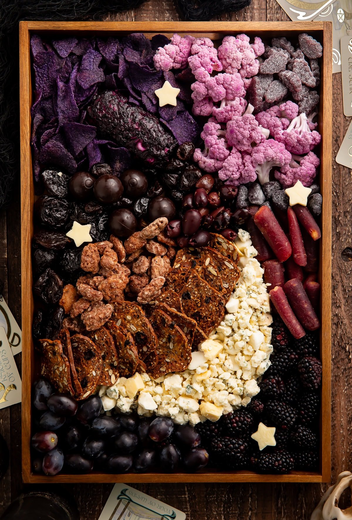 Halloween themed charcuterie board in wooden tray featuring purple and black foods arranged with star cheese cutouts