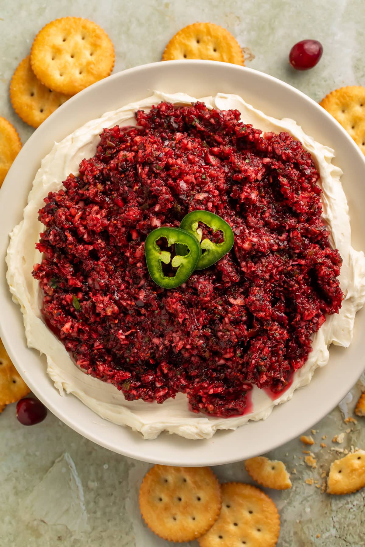 Overhead view of cranberry jalapeño dip on cream cheese in a white bowl, surrounded by Ritz crackers and fresh cranberries on a gray surface.