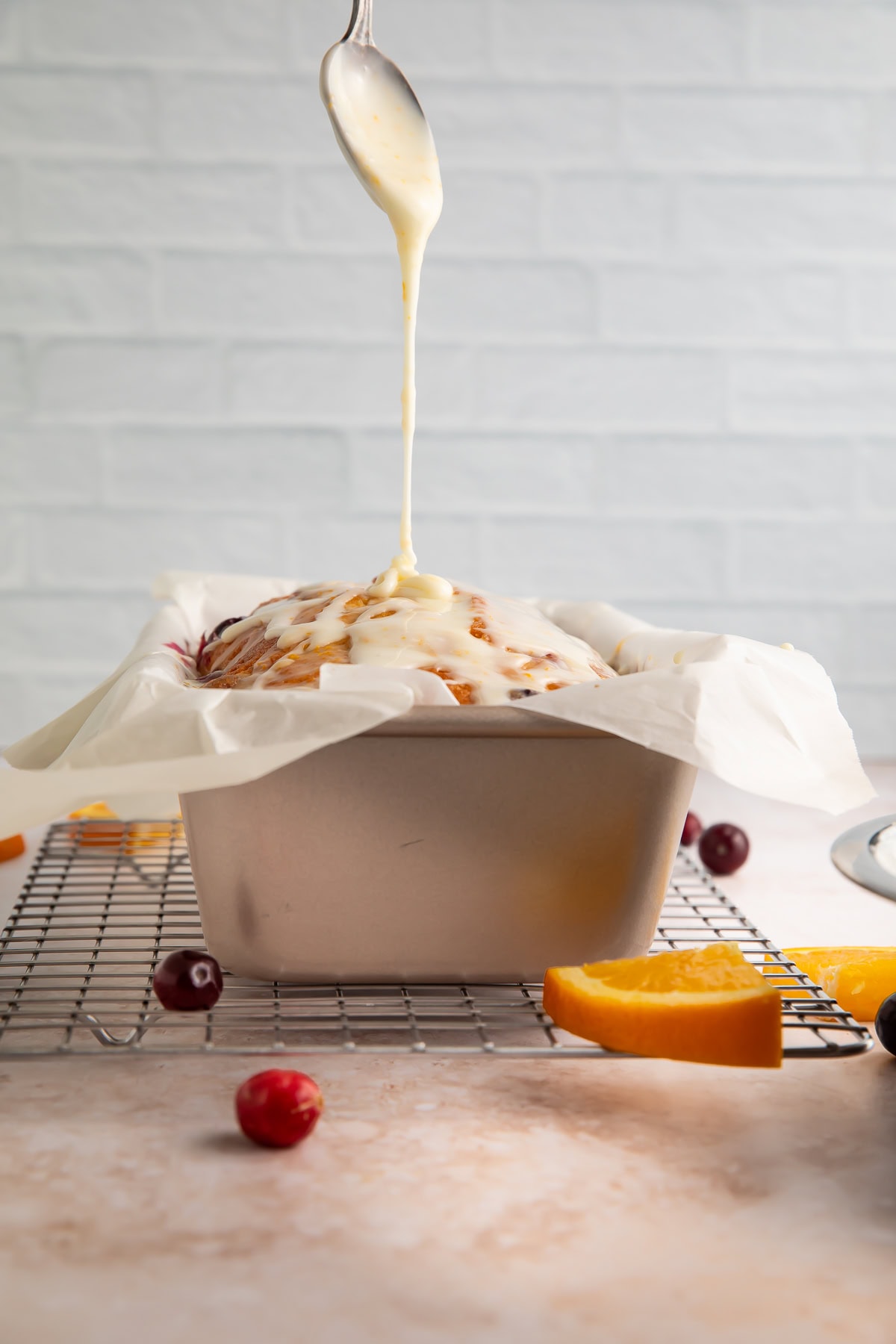 White glaze being drizzled from a spoon onto with fresh cranberries and orange slices nearby
