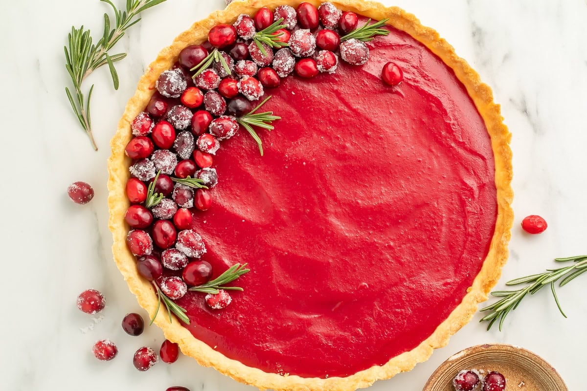 Full cranberry tart on white marble surface with sugared cranberry garnish and fresh rosemary