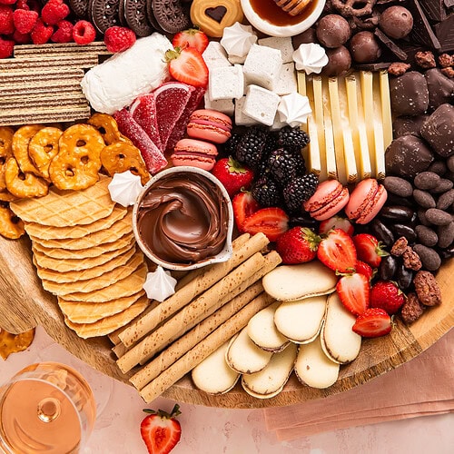 Overhead view of dessert charcuterie board with chocolates, cookies, fresh strawberries, raspberries, blackberries, cheese, honey, pretzels, and waffle cookies on wooden board.
