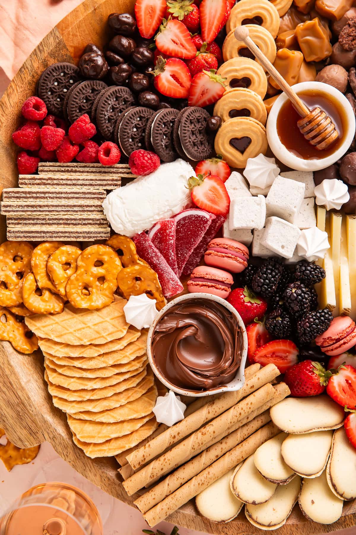 Close-up view of dessert board showing Oreos, waffle cookies, fresh strawberries, raspberries, honey bowl with dipper, chocolate dip, marshmallows, and pink macarons on wooden platter.