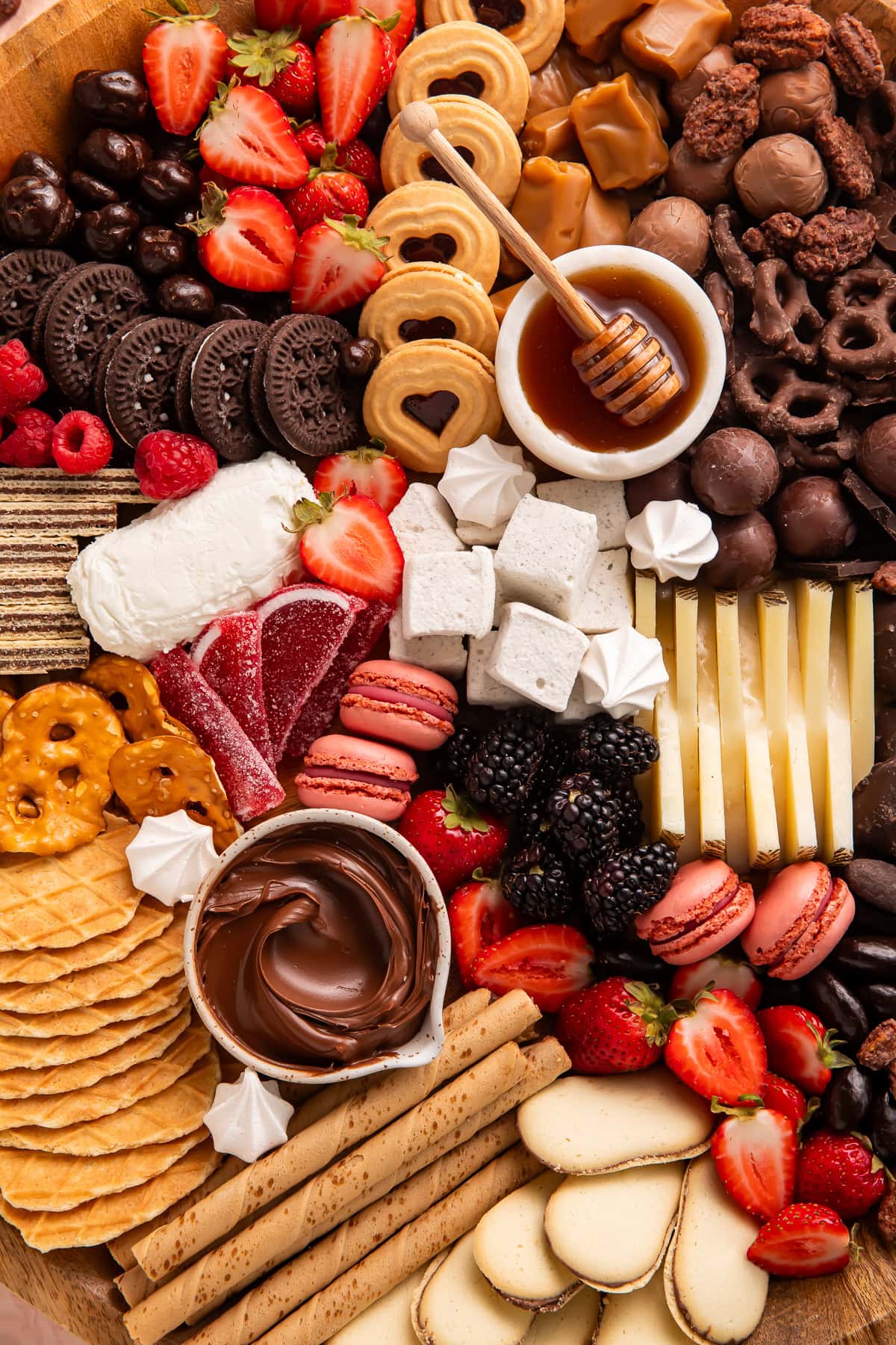 Angled view of complete dessert charcuterie board with chocolate varieties, cookies, fresh berries, cheese, honey bowl, pretzels, macarons, and waffle cookies on round wooden board.