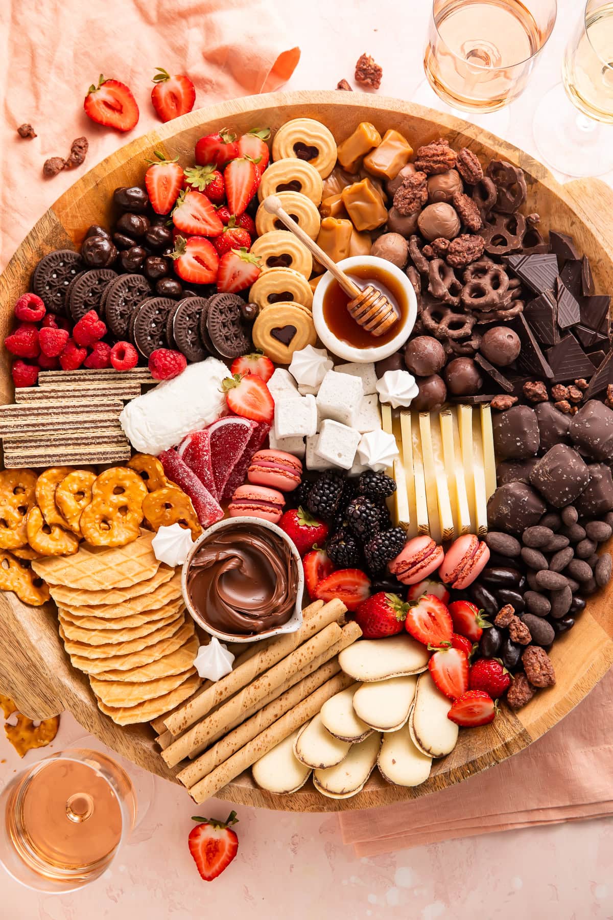 Full dessert charcuterie board on wooden platter with wine glasses and strawberries in background, featuring chocolates, cookies, berries, cheese, honey, and assorted sweets.