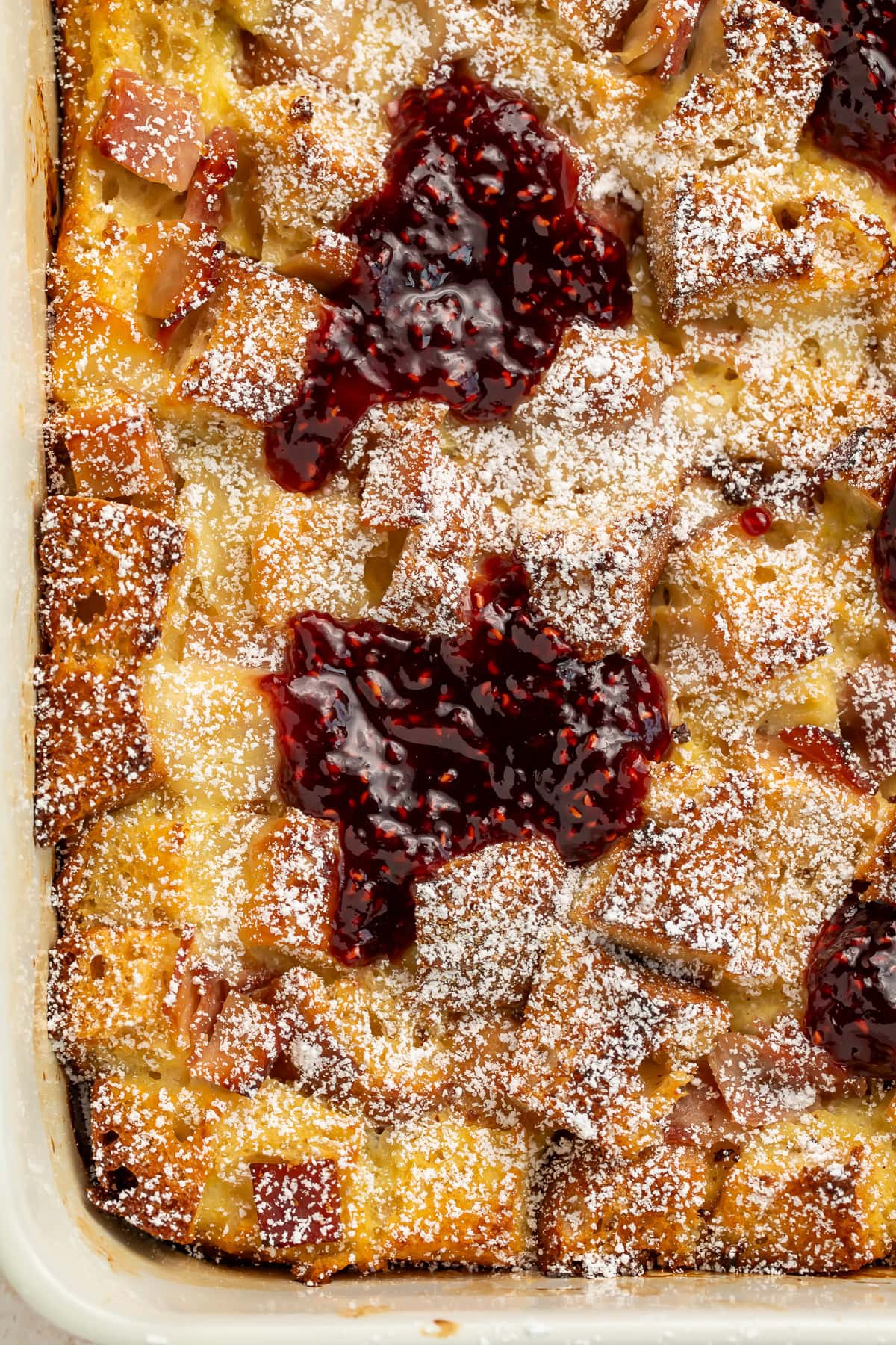 Golden-brown Monte Cristo breakfast casserole topped with powdered sugar and raspberry preserves in a white baking dish.