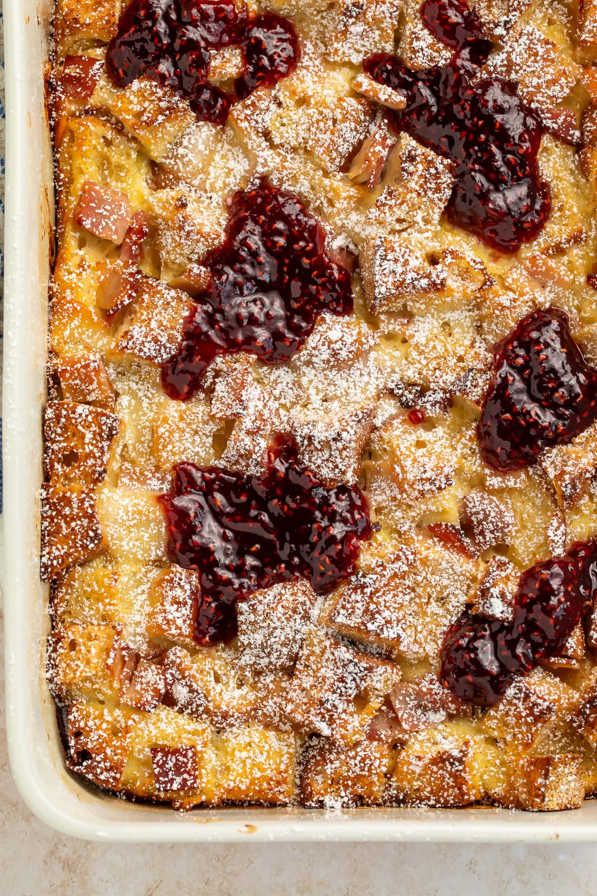 Baked Monte Cristo breakfast casserole in a white pan with powdered sugar and raspberry jam topping.