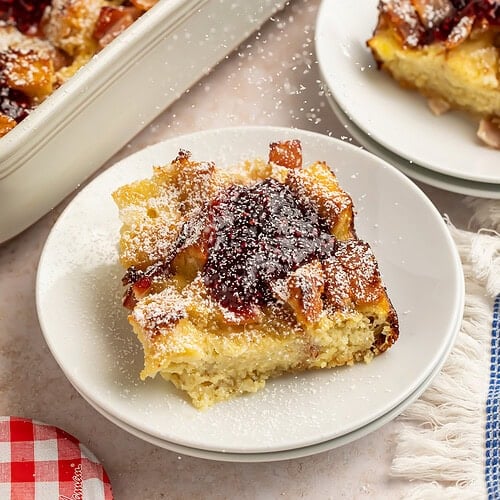 Monte Cristo casserole slices on white plates with raspberry jam and powdered sugar.