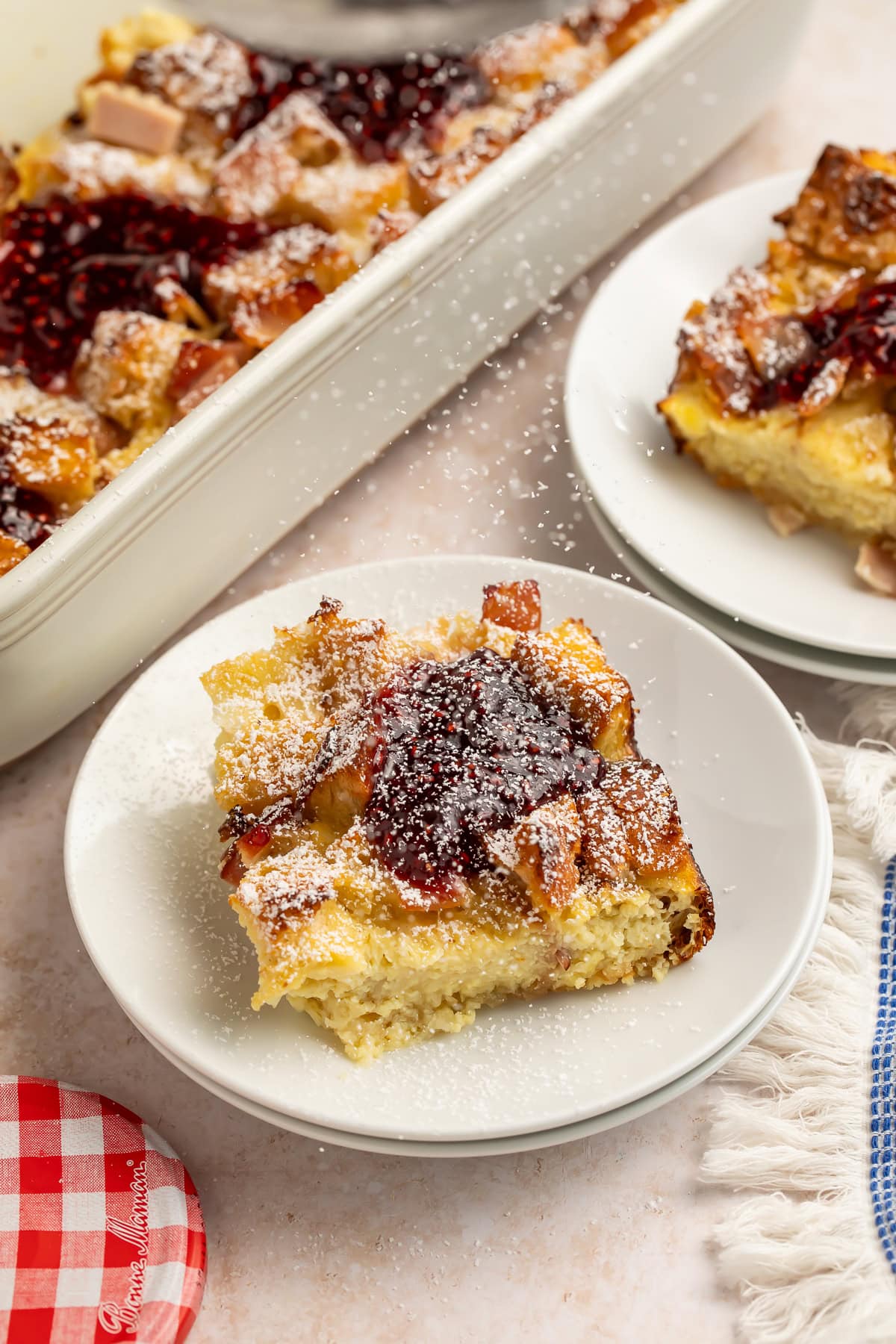 Monte Cristo casserole slices on white plates with raspberry jam and powdered sugar.
