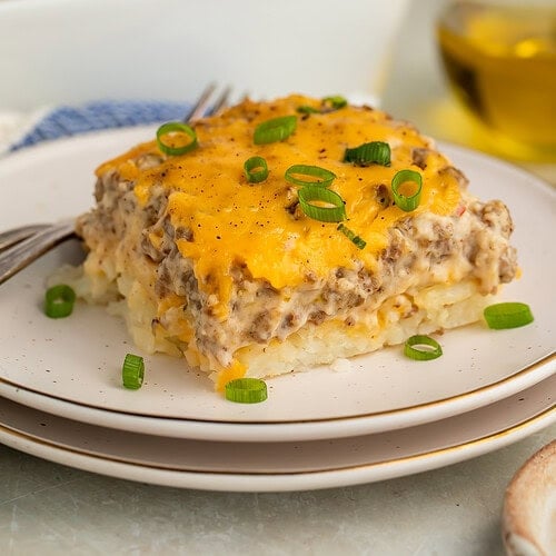 Slice of sausage hashbrown breakfast casserole on white plate showing creamy layers with green onion garnish