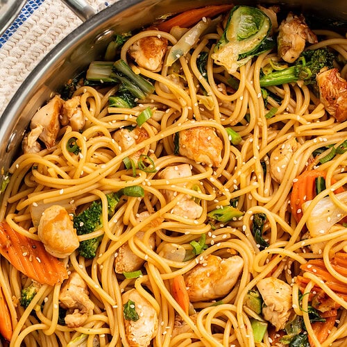 Sesame garlic chicken lo mein in a large skillet with noodles, broccoli, carrots, and chicken topped with sesame seeds