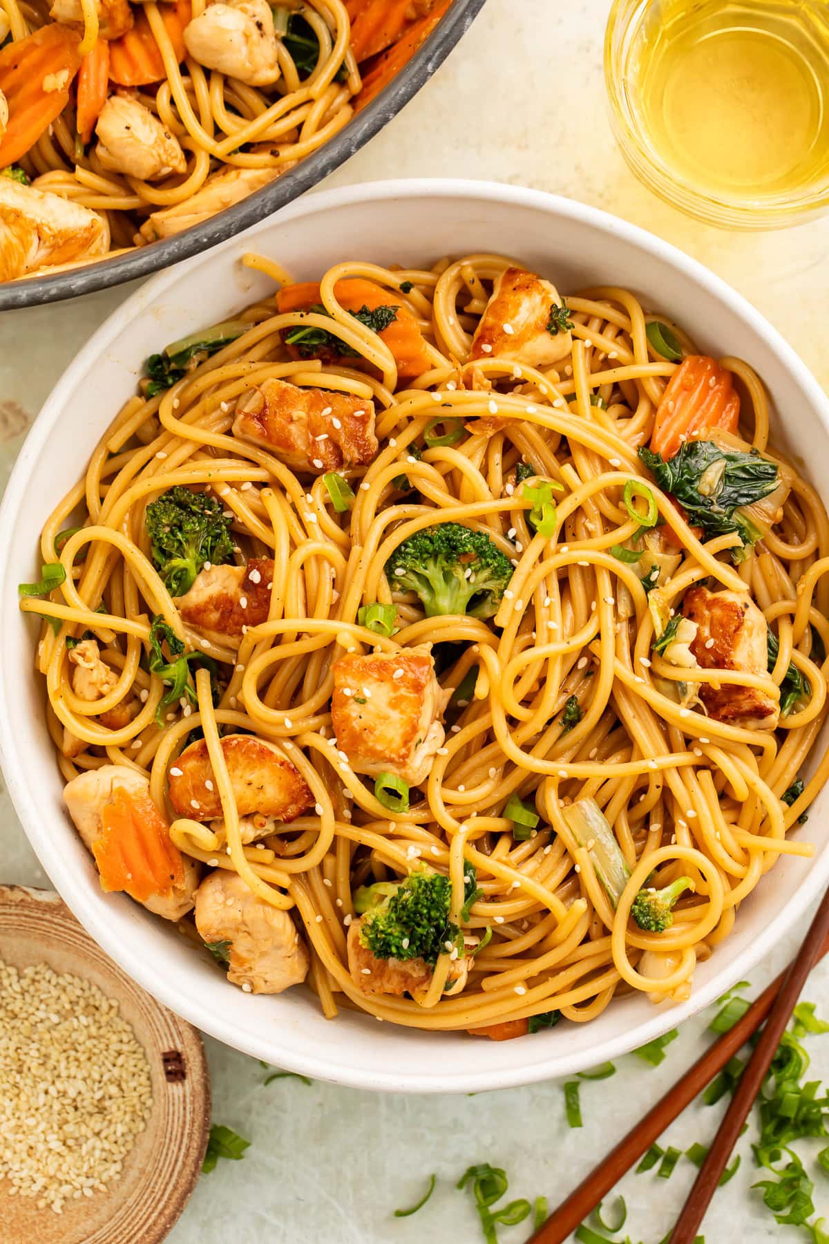 Bowl of sesame garlic chicken lo mein with chopsticks, sesame seeds, and green onions on the side