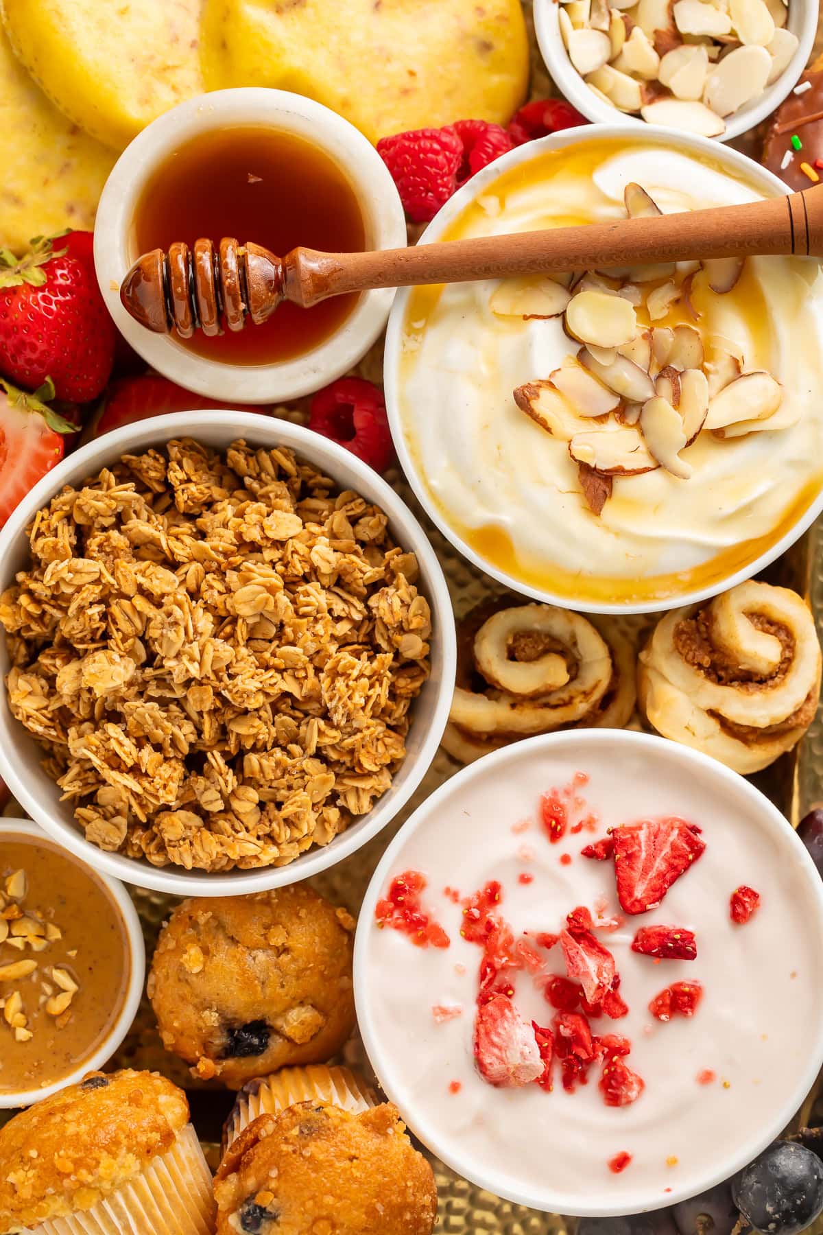 Overhead view of a breakfast charcuterie board with yogurt bowls, granola, fresh berries, waffles, bagels, muffins, and honey on a wooden surface.