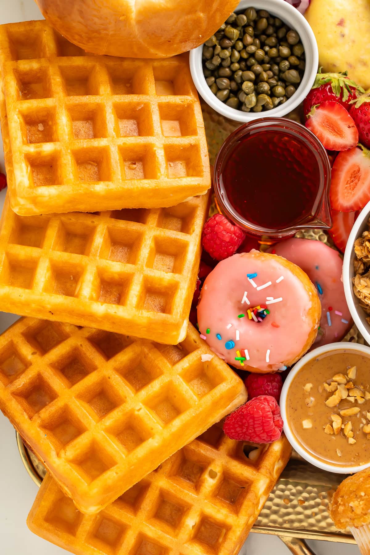 Close-up of golden Belgian waffles, a pink frosted donut, capers, and fresh strawberries on a breakfast charcuterie board.