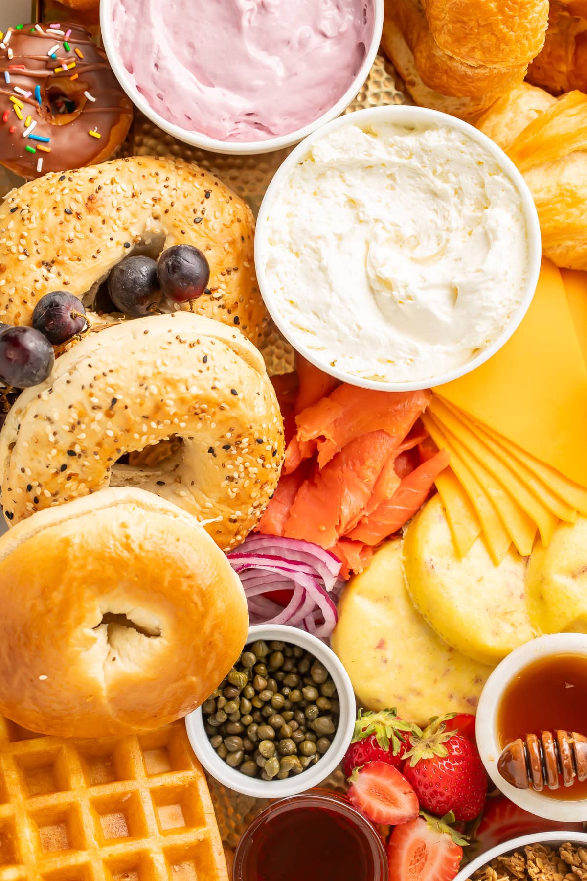 Breakfast charcuterie board with everything bagels, plain bagels, cream cheese, smoked salmon, capers, cheese slices, waffles, and fresh fruit.