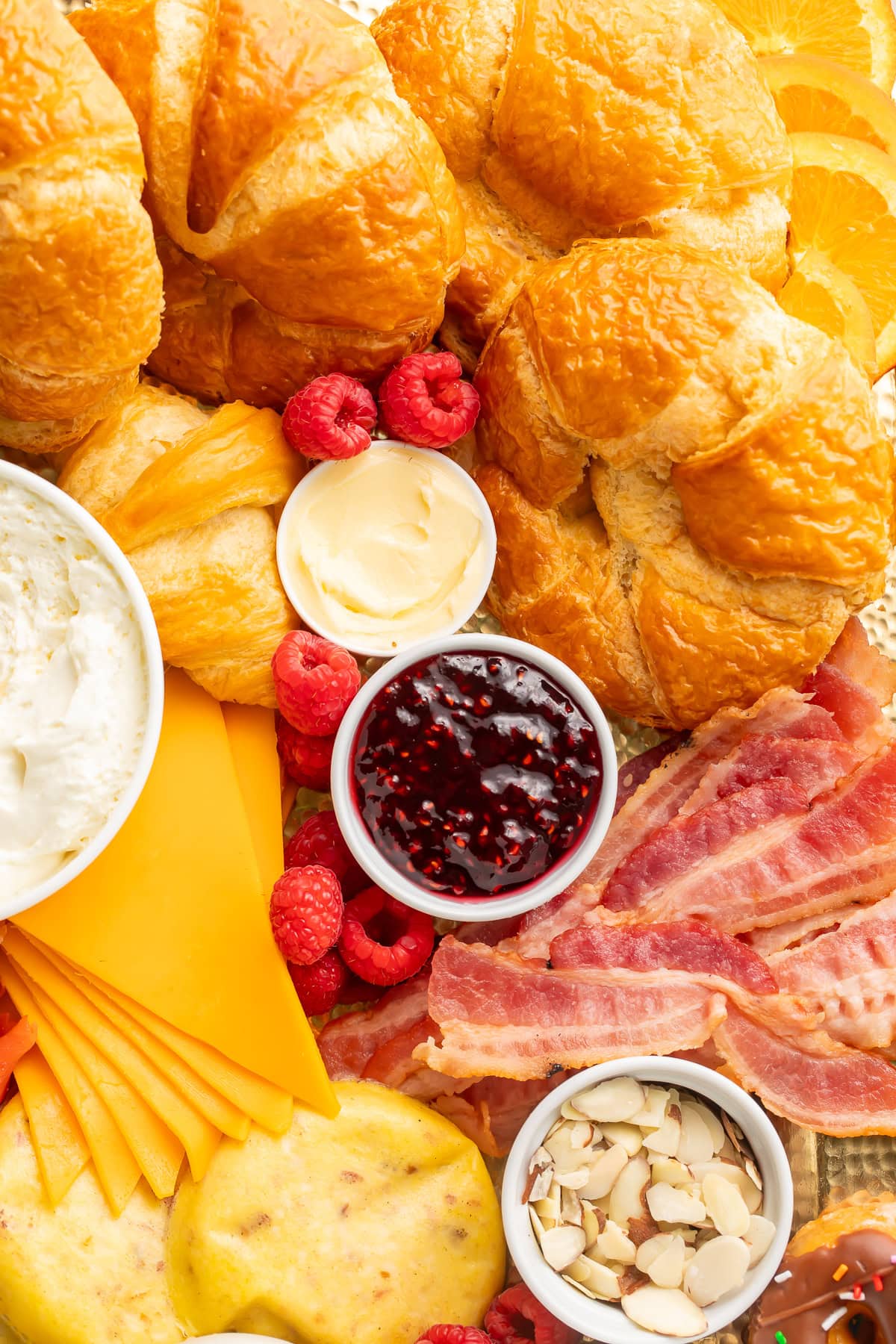 Close-up of flaky golden croissants, bacon strips, cream cheese, jam, cheese slices, and fresh raspberries on a breakfast charcuterie board.