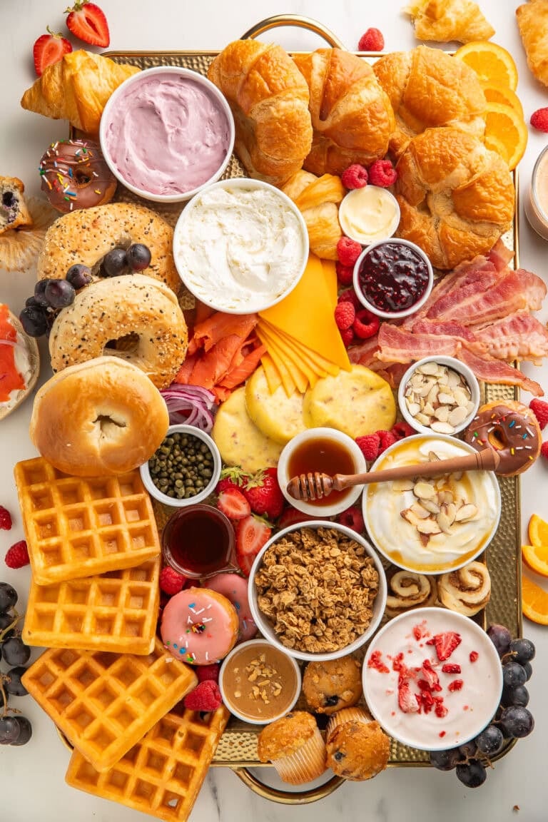 Full breakfast charcuterie board on a wooden tray with bagels, croissants, waffles, yogurt, bacon, cheese, fruit, muffins, and donuts.