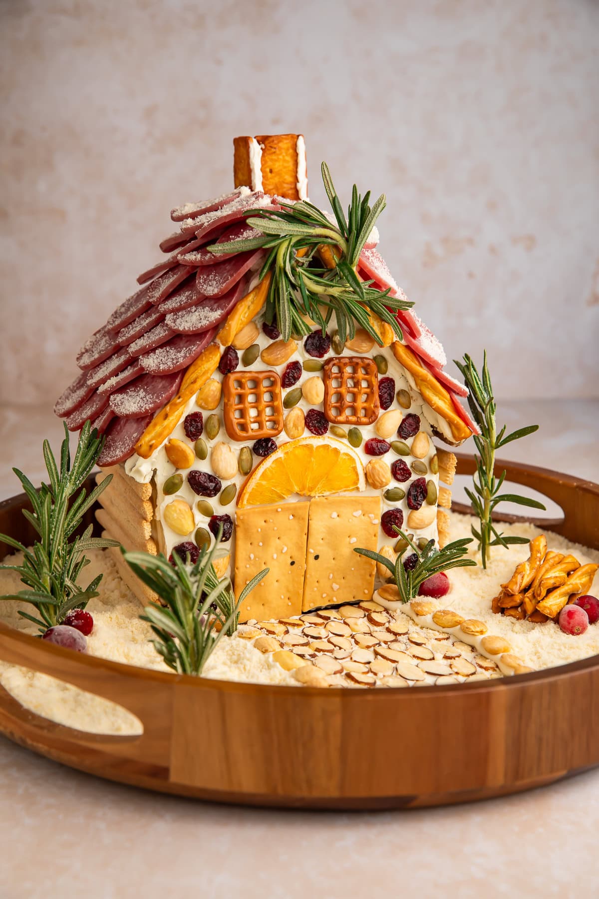 Savory charcuterie chalet with salami shingle roof, pretzel chimney, nuts, and parmesan cheese snow on wooden serving board.