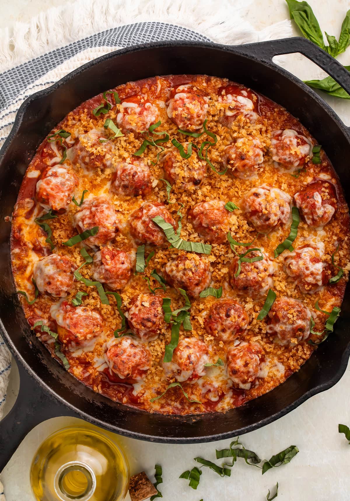 Full cast iron skillet of chicken parmesan meatballs with golden breadcrumbs, fresh basil, and olive oil on white countertop.