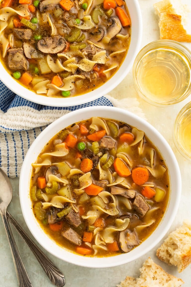 Two white bowls filled with beef soup containing egg noodles, carrots, mushrooms, and vegetables on blue textured placemat.