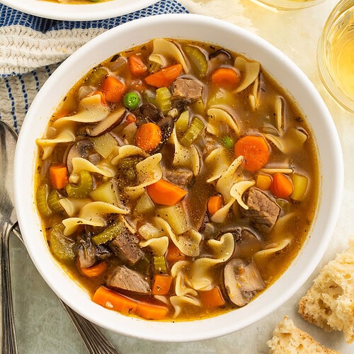 Close-up of white bowl with beef noodle soup showing chunks of meat, wide noodles, sliced carrots, and broth.